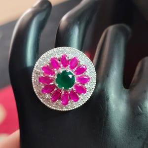 May include: A silver-toned ring featuring a central round green gemstone surrounded by a halo of oval-shaped vibrant pink stones. The outer edge of the circular setting is encrusted with small, clear, sparkling white stones in a pavé style.