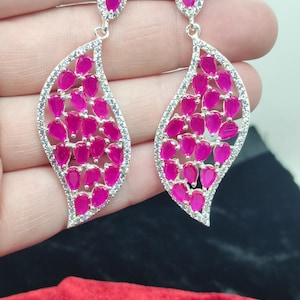 Peut inclure: Une paire de boucles d'oreilles pendantes argentées avec une monture en forme de feuille incrustée de petites pierres claires. L'intérieur de chaque boucle est orné de multiples pierres rose vif en forme de goutte et d'un clou rose.