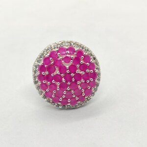 May include: A circular ring featuring a cluster of round, vibrant pink stones set in a silver-toned metal frame. The central pink stones are surrounded by a border of small, clear, sparkling white stones, all held in place by metal prongs.