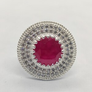 May include: A circular silver ring featuring a large, round, deep red centre stone. The stone is surrounded by two concentric halos of small, clear, sparkling white stones set in a textured silver frame with a beaded edge detail.
