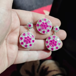 May include: A jewellery set consisting of two circular drop earrings and one matching circular pendant. Each piece features a central cluster of round, vibrant pink stones surrounded by a border of small, clear, sparkling white stones.