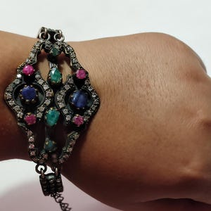 May include: A dark metal bracelet featuring an ornate, elongated central design encrusted with small clear stones. The piece is accented with larger, round-cut blue, green, and pink gemstones arranged symmetrically within the metal framework.