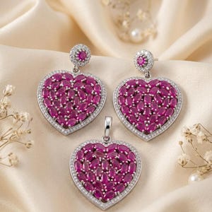Peut inclure: Un ensemble de bijoux composé de deux boucles d'oreilles pendantes en forme de cœur et d'un pendentif assorti. Chaque pièce présente un amas de pierres fuchsia taille marquise entouré d'un bord de petites pierres blanches serties en métal argenté.