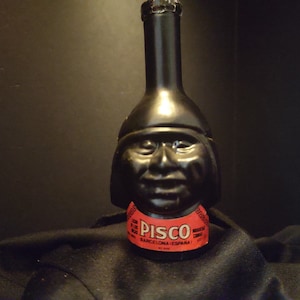 May include: A black glass bottle shaped like a person's head with a red label that says "Pisco Barcelona Espana".