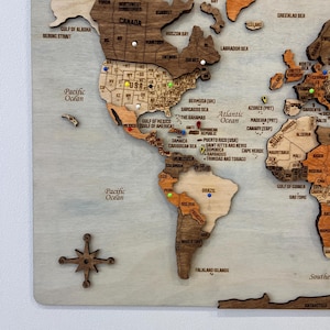 May include: A multi-layered wooden world map wall art piece featuring laser-cut continents in varying shades of brown. The map includes engraved country names, state abbreviations, and a compass rose, accented with colorful push pins.