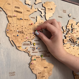 Puede incluir: Un mapa del mundo de madera con contornos de países y fronteras estatales, con alfileres de colores que marcan ubicaciones. Una mano coloca un alfiler verde en el mapa. El mapa incluye el texto "USA" y "CANADA".