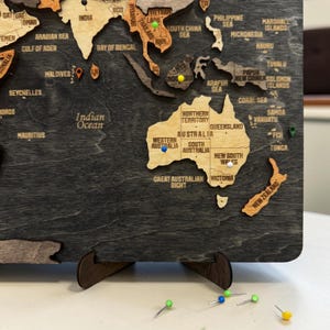 May include: A wooden world map wall art piece featuring a dark gray ocean background and light wood continents. The map includes engraved text for countries and regions, with colorful push pins inserted into various locations on the board.