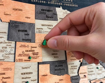 May include: A wooden USA National Parks map featuring laser-cut states in varying shades of wood grain. The map includes text labels for states, cities, and parks, with small green tree-shaped markers placed on specific locations.