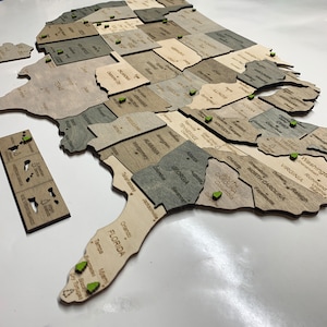 May include: A wooden United States map puzzle with individual state pieces in varying shades of gray and tan. Each state is labeled with its name. Green pins mark cities. The U.S. Virgin Islands and American Samoa are also included.