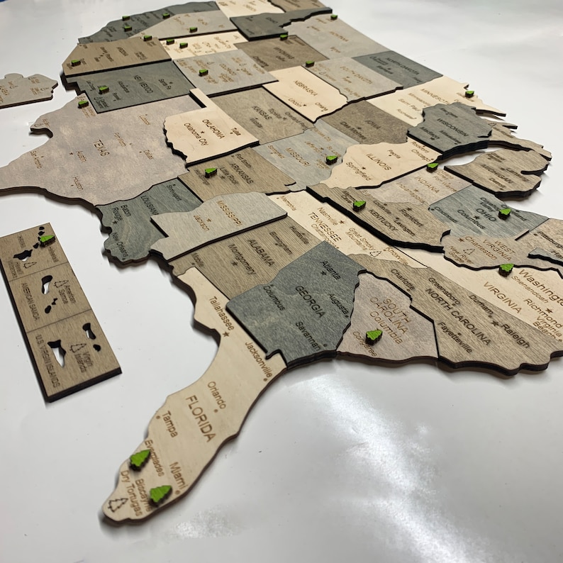 May include: A wooden United States map puzzle with individual state pieces in varying shades of gray and tan. Each state is labeled with its name. Green pins mark cities. The U.S. Virgin Islands and American Samoa are also included.