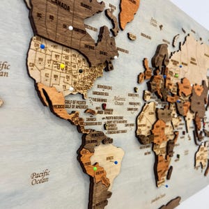 May include: A multi-layered wooden world map wall art piece featuring laser-cut continents and countries in varying shades of brown. The map includes engraved text for country names, states, and oceans, accented with colorful push pins.