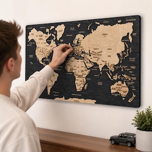 May include: A rectangular wall-mounted world map featuring light-colored wooden continents set against a dark, textured black background. The map displays country names and borders, with small pins inserted into various locations across the landmasses.