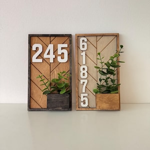 Op de afbeelding: Twee rechthoekige houten wandborden met witte huisnummers. Het linkerbord toont 245 met een donkere lijst en plantenbak. Het rechterbord toont 61875 met een lichte lijst en plantenbak. Beide zijn voorzien van groen kunstmatig loof.
