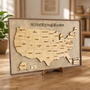 Può includere: Mappa in legno degli Stati Uniti con contorni degli stati e puntine. La mappa è montata su una cornice di legno e include il testo "United States of America". La mappa è esposta su un supporto in legno.