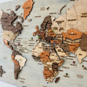 Pode incluir: Um mapa-múndi de madeira multicamadas para parede com vários tons de veios de madeira castanhos e bronzeados. Os continentes estão em relevo, exibindo nomes de países e fronteiras gravados, acentuados com pequenos percevejos coloridos.