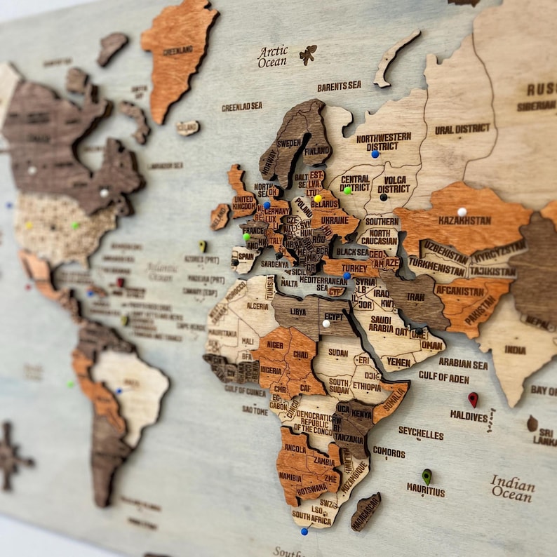 May include: A multi-layered wooden world map wall art piece featuring various shades of brown and tan wood. The map includes engraved country names, regional labels, and ocean names, accented with small, colorful push pins placed in various locations.