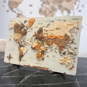 May include: A multi-layered wooden world map wall art piece featuring various shades of brown and tan wood tones. The map includes engraved country names, ocean labels, and small colorful push pins marking specific locations across the continents.