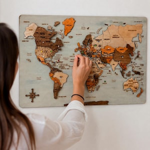 May include: A multi-layered wooden world map wall art piece featuring various shades of brown and tan wood grain. The map includes engraved country names, ocean labels, and a decorative compass rose in the bottom left corner.