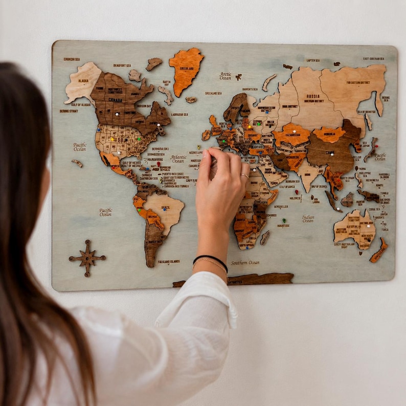 May include: A multi-layered wooden world map wall art piece featuring various shades of brown and tan wood grain. The map includes engraved country names, ocean labels, and a decorative compass rose in the bottom left corner.