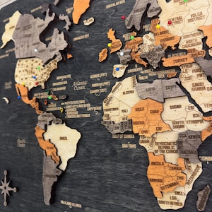 May include: A multi-layered wooden world map wall art piece featuring a dark charcoal background with continents in varying shades of light wood, tan, and orange. The map includes engraved country names and colorful push pins.