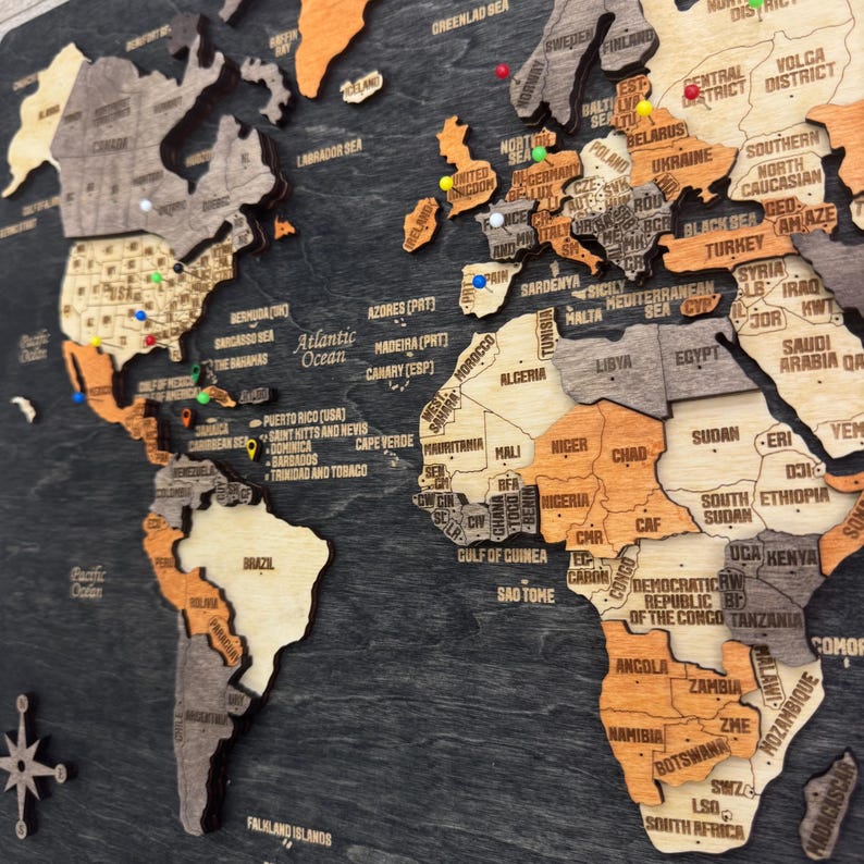 May include: A multi-layered wooden world map wall art piece featuring a dark charcoal background with continents in varying shades of light wood, tan, and orange. The map includes engraved country names and colorful push pins.