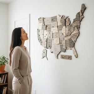 May include: A wooden map of the United States is mounted on a white wall. The map is made of various shades of gray and beige wood, with each state labeled. Small pins mark locations. A person is looking up at the map.