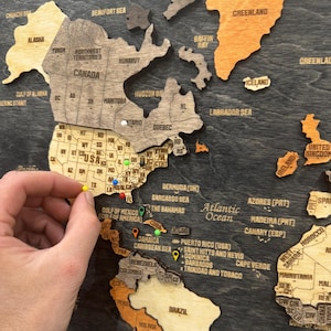 May include: A multi-layered wooden wall map featuring North and South America with engraved country names, state borders, and labels. The map includes various colored push pins inserted into specific locations across the USA and Caribbean regions.