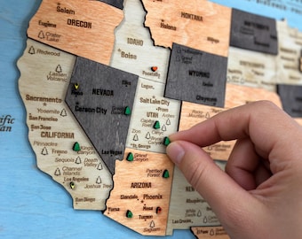 Pode incluir: Um mapa de madeira dos parques nacionais dos EUA com formas de estados cortadas a laser em vários tons de madeira. O mapa mostra nomes de estados, cidades e parques com ícones. Uma mão coloca um pino verde em forma de árvore em Utah.