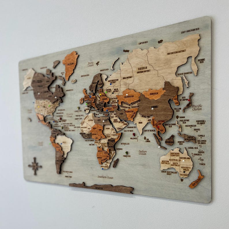 May include: A rectangular wooden world map wall art piece featuring laser-cut continents in varying shades of brown and tan wood. The map includes engraved country names, regional labels, and colorful push pins marking specific locations.