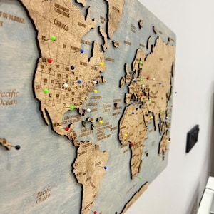 Puede incluir: Mapa del mundo de madera con continentes y nombres de países grabados. El mapa está montado sobre un fondo azul claro y presenta chinchetas de colores que marcan ubicaciones. El mapa incluye los océanos Pacífico y Atlántico.