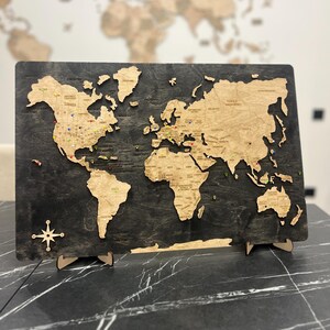 Puede incluir: Un mapa del mundo de madera con un fondo gris oscuro y continentes de color marrón claro. El mapa está detallado con los nombres de los países y las ubicaciones de los alfileres. Una rosa de los vientos está en la esquina inferior izquierda. El mapa está soportado por soportes de madera.
