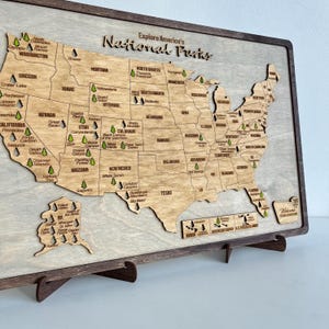 Può includere: Mappa in legno degli Stati Uniti con i contorni degli stati e le posizioni dei parchi nazionali. La mappa è marrone chiaro con una cornice marrone scuro e il testo "Explore America's National Parks."
