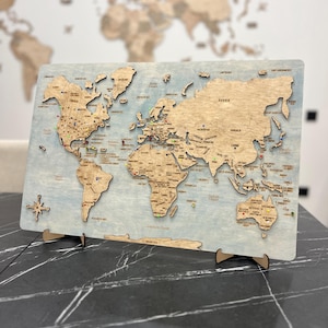 Pode incluir: Um mapa-múndi retangular de madeira montado sobre um painel pintado de azul claro. Os continentes são cortados a laser num tom de madeira natural, com nomes de países gravados, uma rosa dos ventos e pequenos percevejos coloridos.
