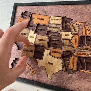 Può includere: Una mappa da parete in legno degli Stati Uniti con pezzi individuali per ogni stato in varie tonalità di marrone. La mappa è incorniciata in nero e ogni stato è inciso al laser con il proprio nome. Un pezzo del New Mexico viene inserito nella mappa.