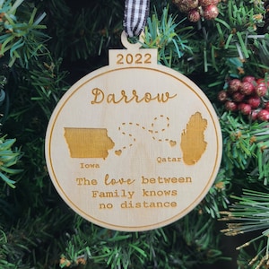 Op de afbeelding: Een houten kerstversiering met de tekst "Darrow" en "2022" erop. De versiering heeft ook de contouren van Iowa en Qatar met een stippellijn die ze verbindt. De tekst "De liefde tussen familie kent geen afstand" staat ook op de versiering.