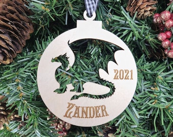 May include: A wooden Christmas ornament shaped like a circle with a dragon silhouette cut out. The ornament is painted white and has the name "Zander" and the year "2021" engraved on it.