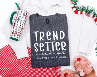 May include: A dark gray t-shirt with white text reading TREND SETTER mockups Real People, Real Products, placed on a white waffle-knit sweater, red and white checkered pants, a nutcracker-patterned tumbler, and fuzzy slippers.