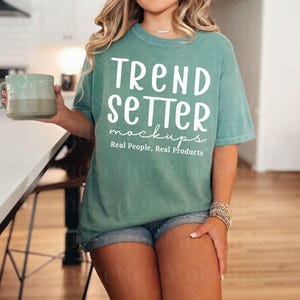 Pode incluir: Uma camiseta verde-azulado com o texto "TREND SETTER mockups Real People, Real Products" em branco. A camiseta é combinada com shorts jeans. Uma pessoa está segurando uma caneca.