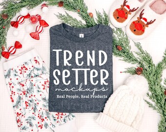 May include: A dark heather gray t-shirt featuring the white text TREND SETTER mockups Real People, Real Products, surrounded by white and red candy ornaments, white reindeer slippers, and a white knit beanie on a pine branch background.