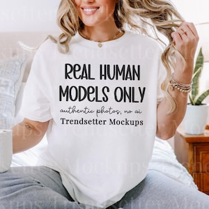 May include: A white t-shirt with the text "REAL HUMAN MODELS ONLY authentic photos, no ai Trendsetter Mockups" in black. The person is wearing gray sweatpants and holding a white mug. The background includes a bed, pillows, and a wooden nightstand.