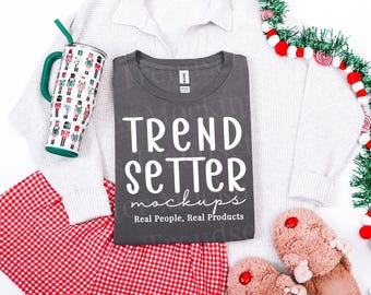 May include: A dark gray t-shirt with white text that reads TREND SETTER mockups Real People, Real Products. The shirt is displayed with a white waffle-knit sweater, red and white checkered shorts, a nutcracker-patterned tumbler, and fuzzy slippers.