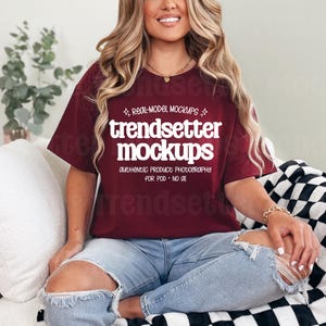 May include: A maroon t-shirt with the text "trendsetter mockups" in white. The shirt is worn with ripped blue jeans. The model is sitting on a white couch with a black and white checkered blanket.