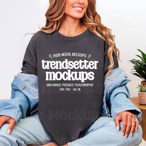 May include: A dark gray long-sleeve t-shirt with the text "trendsetter mockups" in white. The shirt is worn with a light blue denim jacket and light blue jeans. The image includes potted plants on a wooden table.