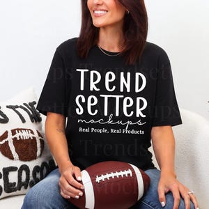 Puede incluir: Camiseta negra con el texto "TREND SETTER mockups Real People, Real Products" en blanco. Un balón de fútbol marrón y blanco se sostiene en el regazo. Un cojín decorativo con las palabras "THIS IS SEASON" está en el fondo.