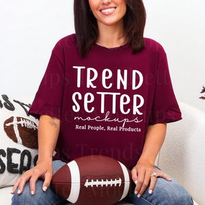 Puede incluir: Una camiseta granate con el texto "TREND SETTER mockups Real People, Real Products" en blanco. Un balón de fútbol americano marrón y blanco se sostiene en el regazo. Se ve un cojín decorativo con un diseño de fútbol y las palabras "Tis the Season".
