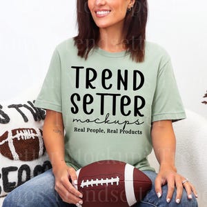 Puede incluir: Una camiseta verde claro con el texto "TREND SETTER mockups Real People, Real Products" en negro. Se sostiene un balón de fútbol americano marrón en el regazo. Un cojín decorativo con un diseño de fútbol y las palabras "Tis the Season" está en el fondo.