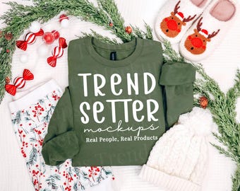 May include: An olive green long-sleeve sweatshirt featuring the white text TREND SETTER mockups Real People, Real Products. The garment is surrounded by holiday-themed items including reindeer slippers, candy ornaments, and a white knit hat.
