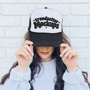 Puede incluir: Gorra de camionero en blanco y negro con el texto "trendsetter Mockups" en una fuente retro audaz. La gorra tiene una visera negra y un panel frontal blanco. La gorra está siendo sostenida por una persona.