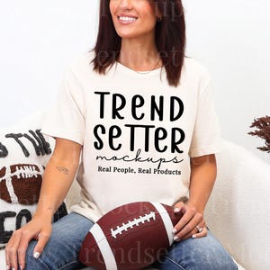 Puede incluir: Una mujer lleva una camiseta color crema con el texto "TREND SETTER mockups Real People, Real Products" en negro. Sostiene un balón de fútbol americano marrón y blanco. Se ve una almohada con un diseño de balón de fútbol.