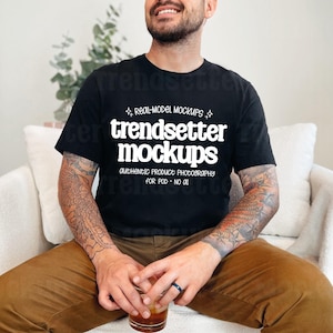 Könnte beinhalten: Schwarzes T-Shirt mit dem Text "trendsetter mockups" in Weiß. Das T-Shirt wird von einer Person getragen, die in einem weißen Stuhl sitzt. Die Person trägt eine braune Hose und hält ein Glas Flüssigkeit. Das Bild ist ein Produkt-Mockup.